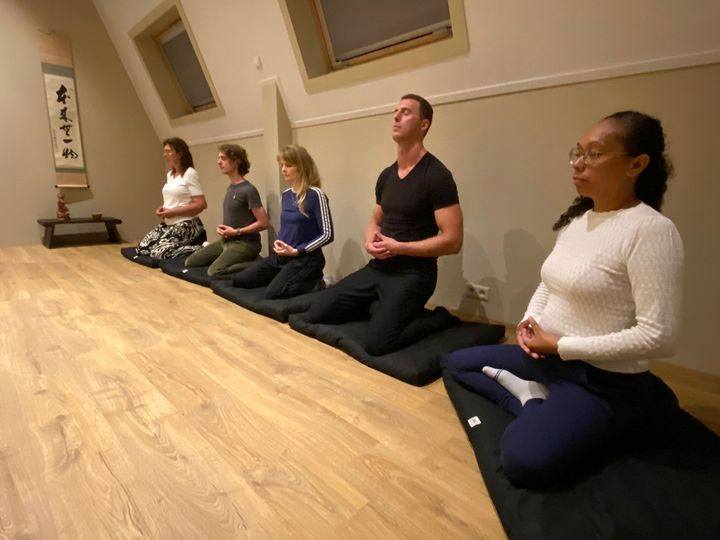 vijf mensen zitten naast elkaar in een rustig ingerichte ruimte op meditatiematten, zenmeditatie