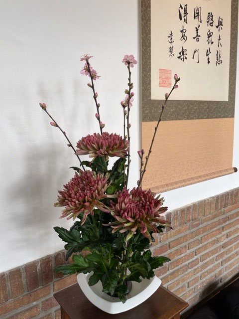 in een lage schaal staat een bloemstuk met chrysanten voor een witte muur met daaraan een Japanse scroll met kanji