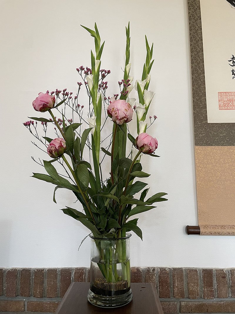 in een glazen vaas staat een bloemstuk met pioenrozen voor een witte muur met daaraan een Japanse scroll met kanji